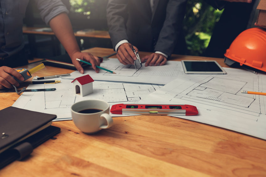 Group of people reviewing architectural plans on a wooden table with a model house, coffee cup, tablet, and hard hat.