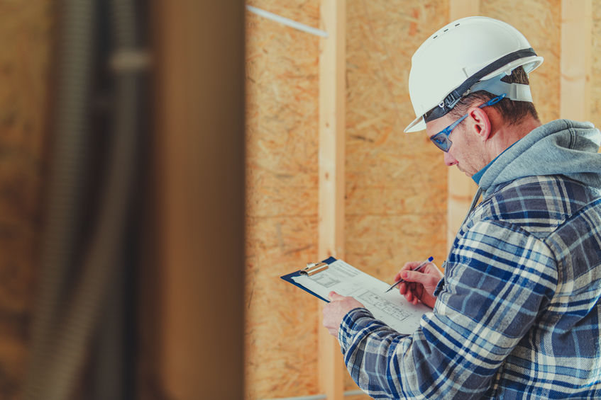 A construction worker wearing a hard hat and safety glasses writes on a clipboard inside a building with wooden walls.