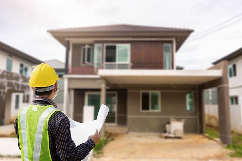 Construction worker in a safety vest and helmet holds blueprints, facing an unfinished two-story house.