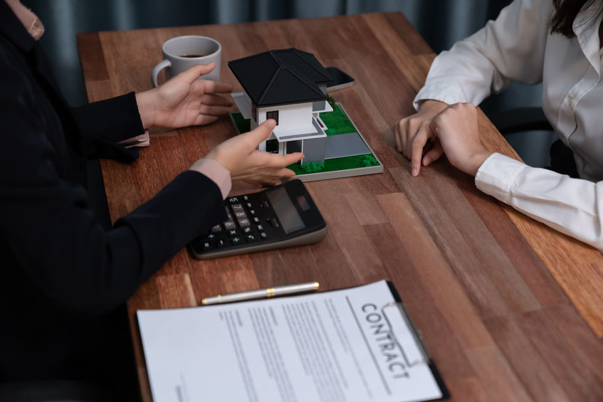 Two people at a table with a house model, discussing a contract. A calculator, pen, and coffee cup are present.